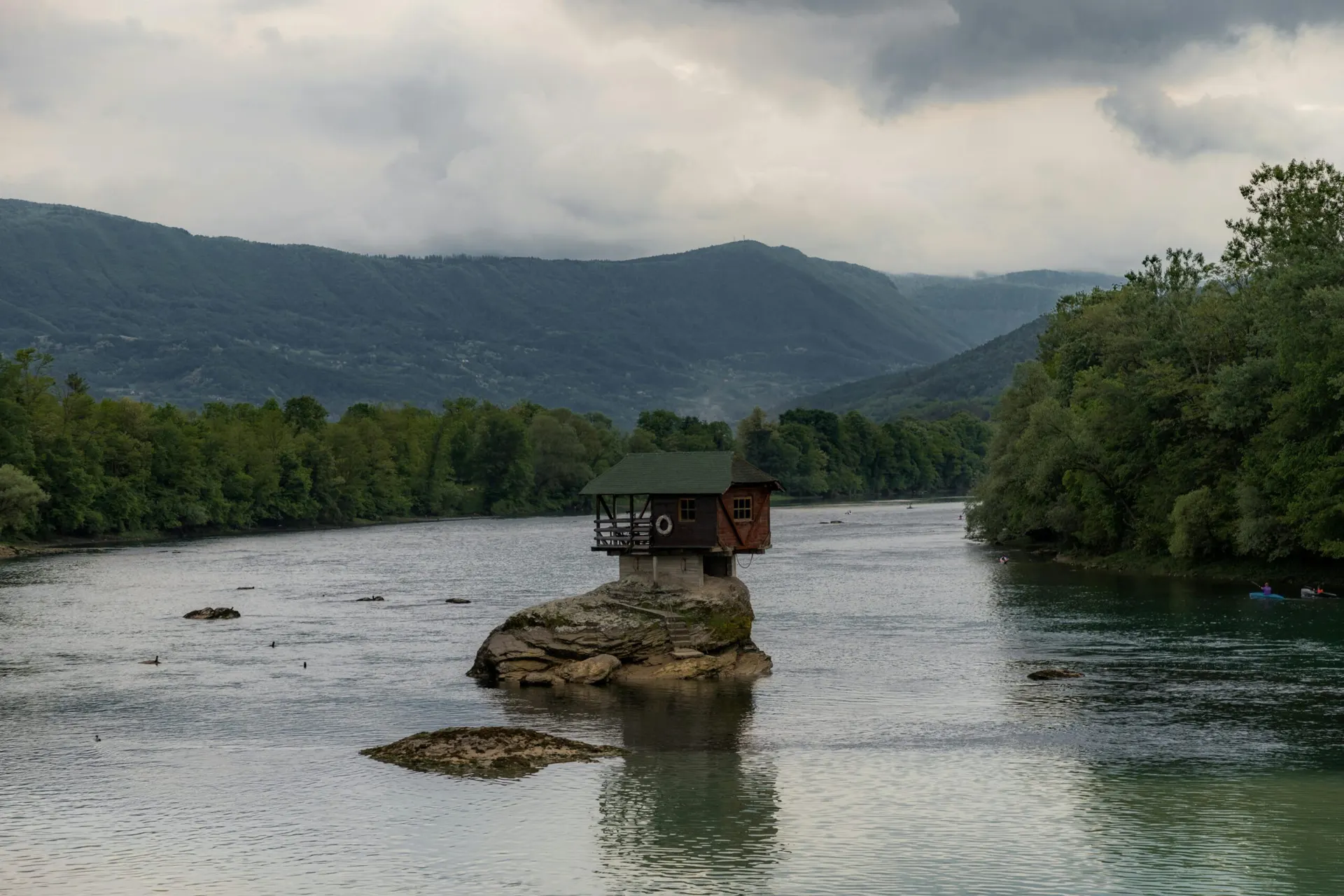Una casetta di legno su uno scoglio in mezzo a un fiume, con le montagne boscose sullo sfondo
